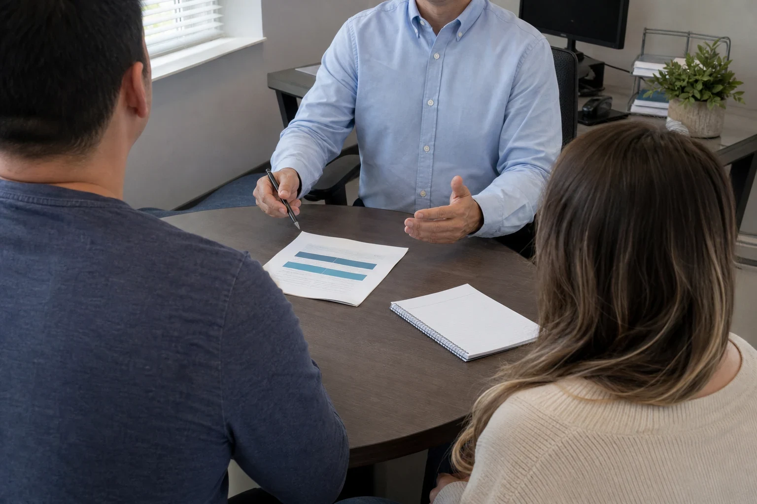 Agent in a light blue button-down shirt gestures while talking with a couple at a round wooden table holding a printed summary and a notepad in a small office